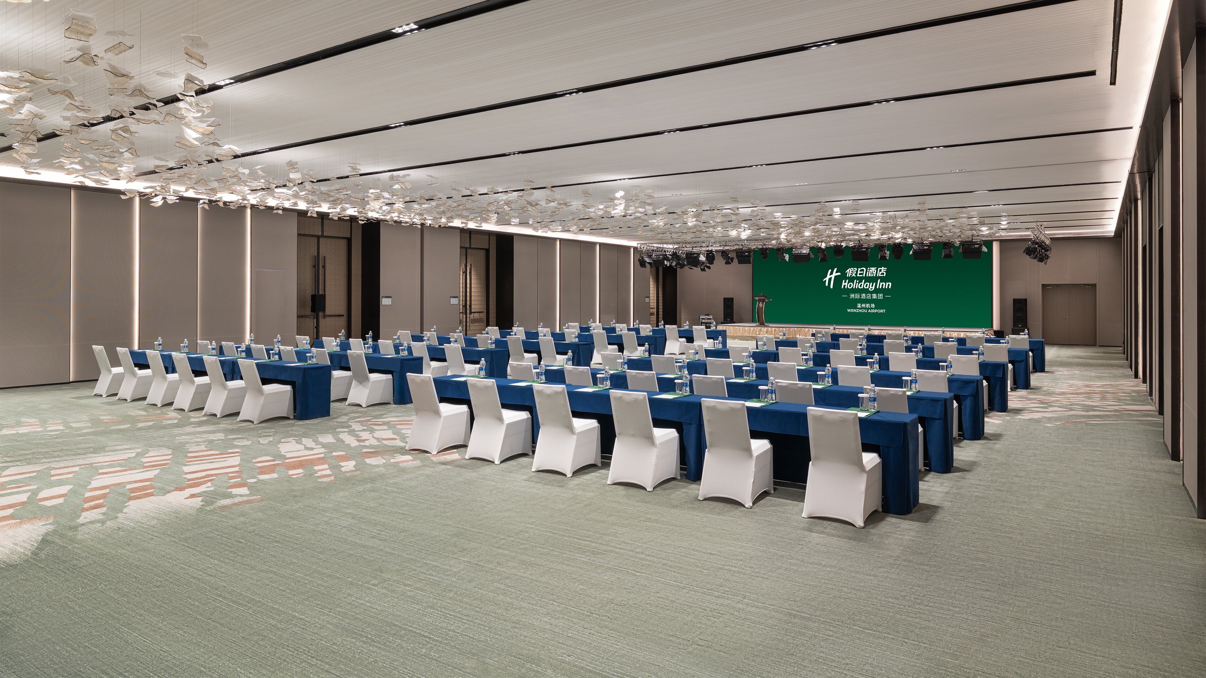 Un salón de conferencias con filas de mesas y sillas, una pantalla verde con texto y una alfombra estampada.
