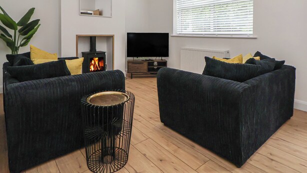 A modern living room with dark sofas, a small side table, a fireplace, a television, and a large window with blinds. - 5 Marsh Lane (Flint)