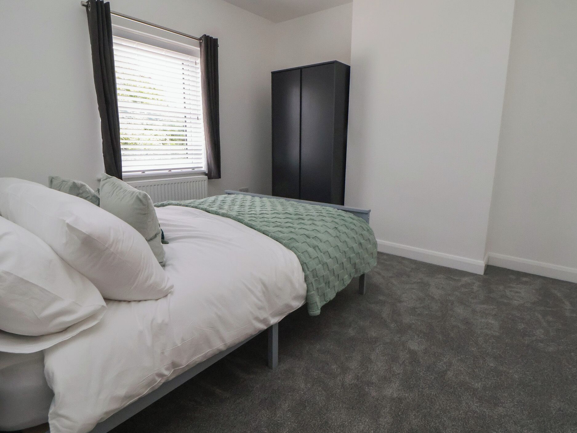 A bedroom with a bed, a radiator, a window with curtains, and a wardrobe.