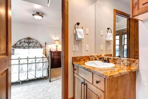 Een slaapkamer met een metalen bedframe, een nachtkastje met een lamp, en een badkamer met een wastafel met granieten aanrechtblad en houten kastjes.