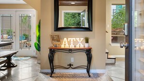 A modern entryway with a wooden console table displaying 'ATX' letters, a mirror above, and a view of the outdoors through a glass door. - ATX Oasis!  3-Story Home w/ Heated Pool, Spa & Sauna - Close to downtown (Austin)
