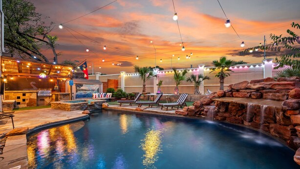 A pool area with a waterfall, lounge chairs, and string lights against a sunset backdrop. - ATX Oasis!  3-Story Home w/ Heated Pool, Spa & Sauna - Close to downtown (Austin)