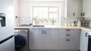 A modern kitchen with grey cabinets, a built-in washing machine, and a window providing natural light. - Lockmans Cottage (Llanymynech)