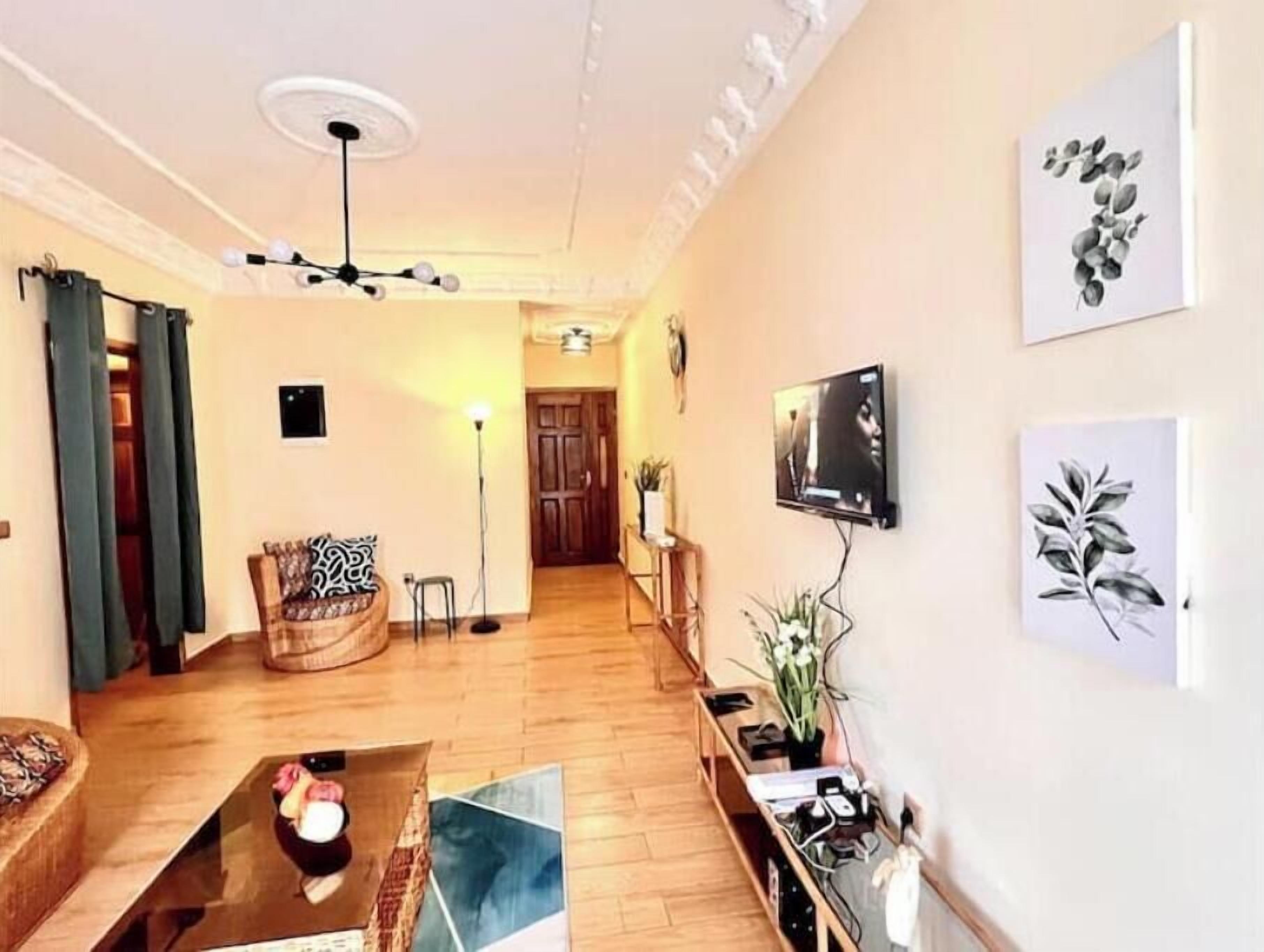 Un salón con un sofá, una mesa de centro, un televisor montado en la pared y una puerta que da a otra habitación.