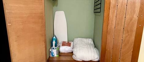 Een kleine, afgesloten ruimte met een houten bankje, een wit kussen, een zak tissues en een hangrek met kleurrijke spullen.