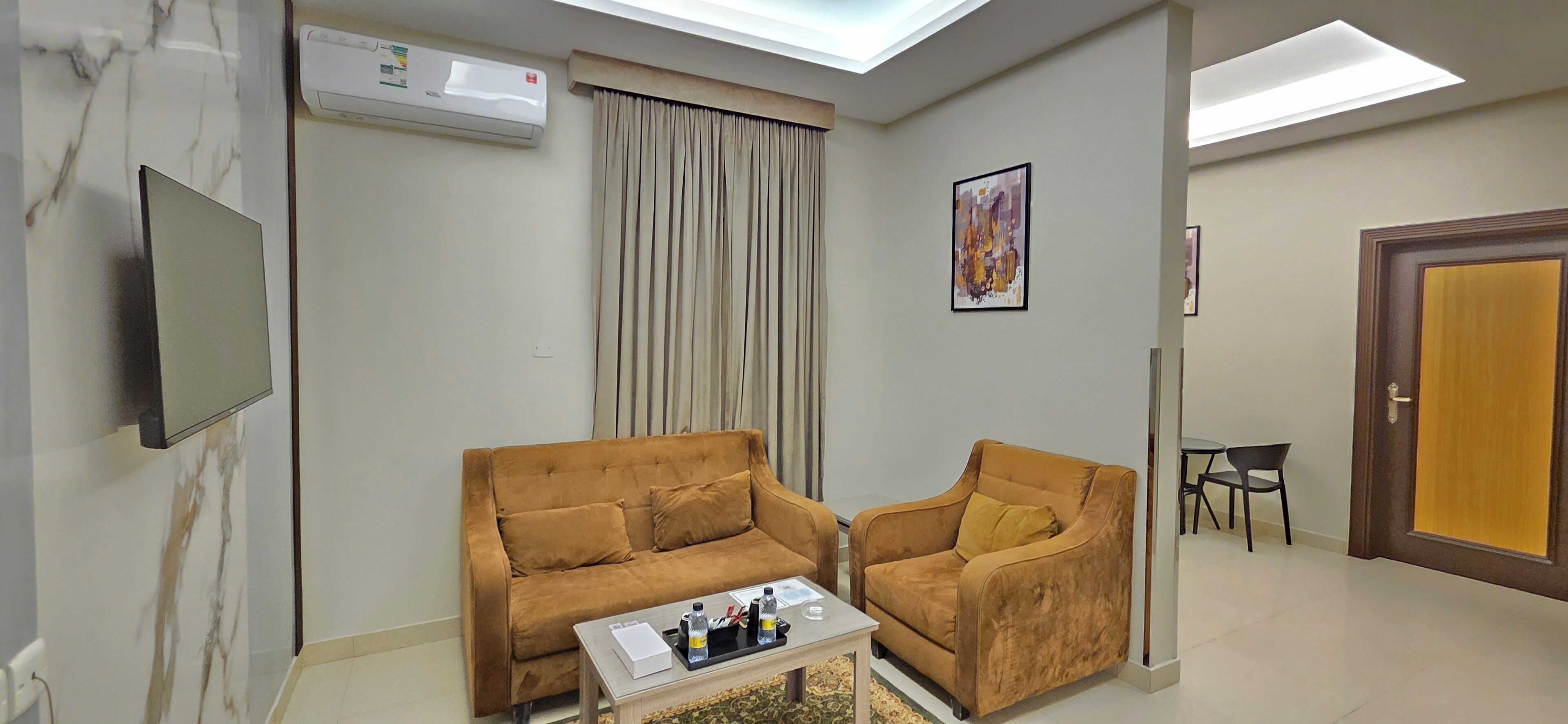 A living room with a sofa, armchair, and a coffee table. There is a flat-screen TV mounted on the wall, an air conditioning unit, and a door with a golden handle.