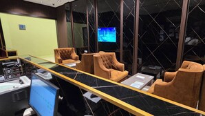 Egy modern szállodai előtér, amelyben található egy recepciós pult, kényelmes ülések, egy nagy képernyős televízió és egy üvegfal.