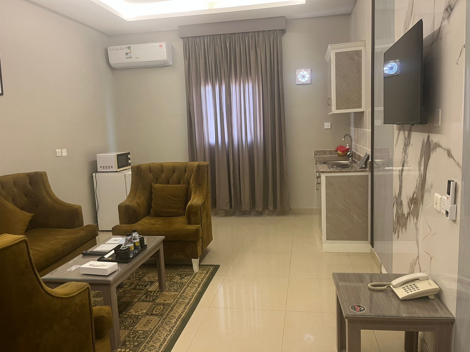 Una sala moderna con sofá, sillón y una mesita de centro. Hay un televisor montado en la pared, una zona de cocina con fregadero y un teléfono sobre una mesita auxiliar.
