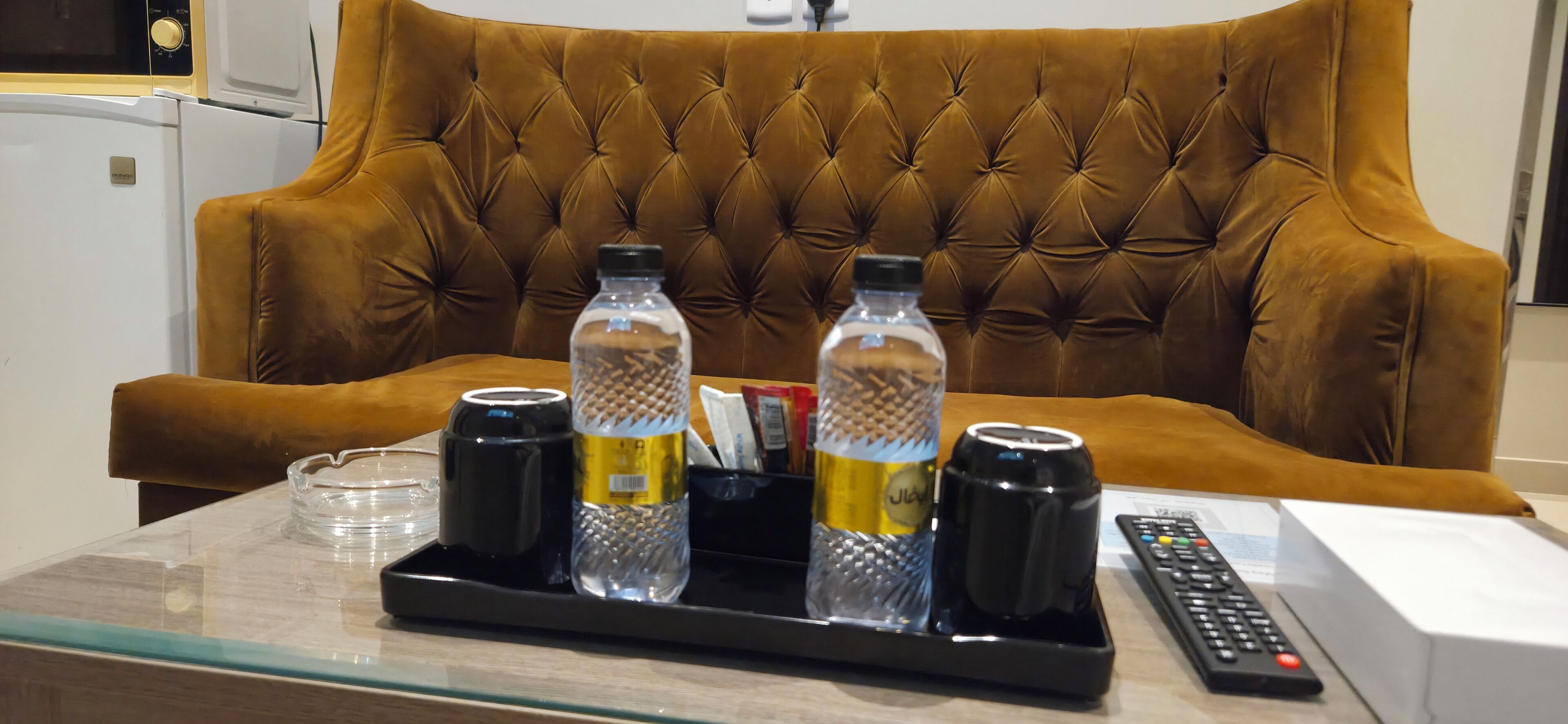 A hotel room with a tufted brown sofa, a tray table with a water bottle, a can, a mug, and a remote control.