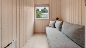 A room with wooden panel walls, a gray sofa with pillows, a small window with a view of greenery and a building, and a radiator on the floor. - Holiday home by Balka Beach with perfect location (Nexø)