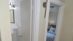 A hallway with a wooden floor leading to a bedroom with a bed and a nightstand. A door on the left leads to a bathroom with a towel rack and a trash can. - Charming 3-bedroom house in delightful Peabody (Peabody)
