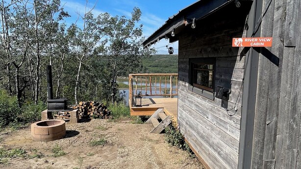 A rustic cabin with a deck overlooking a river, surrounded by trees.