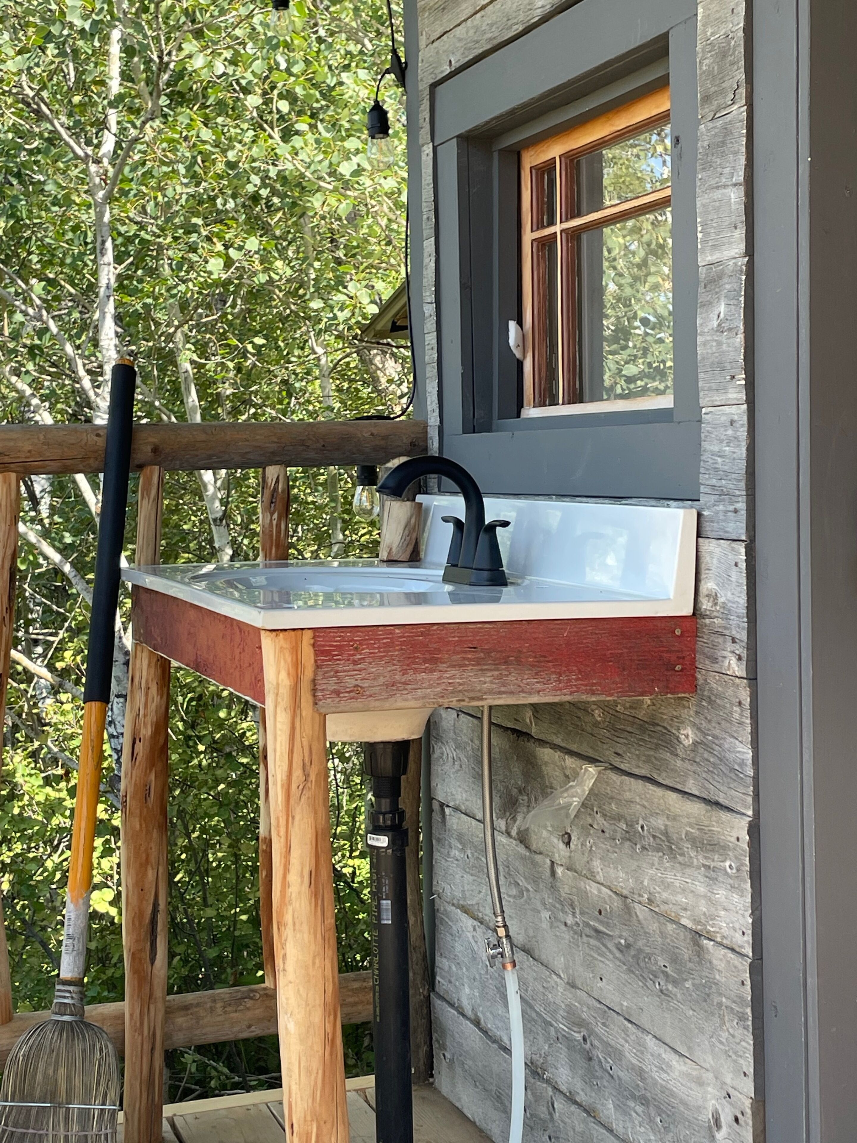 A rustic wooden cabin with a small outdoor sink, a wooden deck, and a window overlooking greenery.