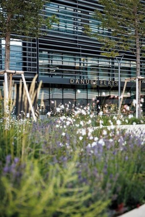 Um edifício moderno com o nome 'DANUBE FLATS' na fachada, rodeado por um jardim com diversas plantas e flores.