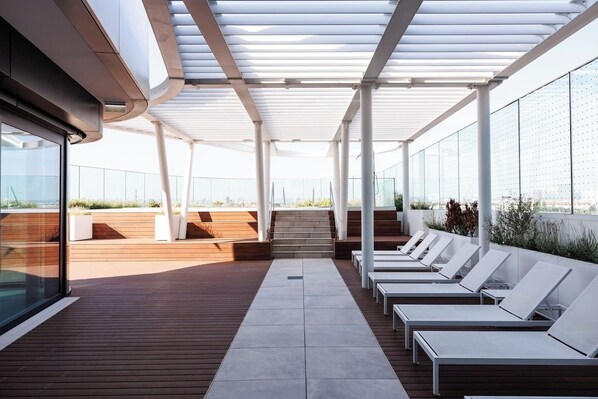A rooftop terrace with wooden flooring, white lounge chairs, and a glass wall offering a city view.
