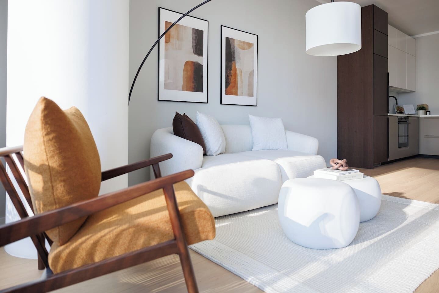 Un salón moderno con sofá, reposapiés y sillón, una cocina al fondo y dos cuadros abstractos en la pared.