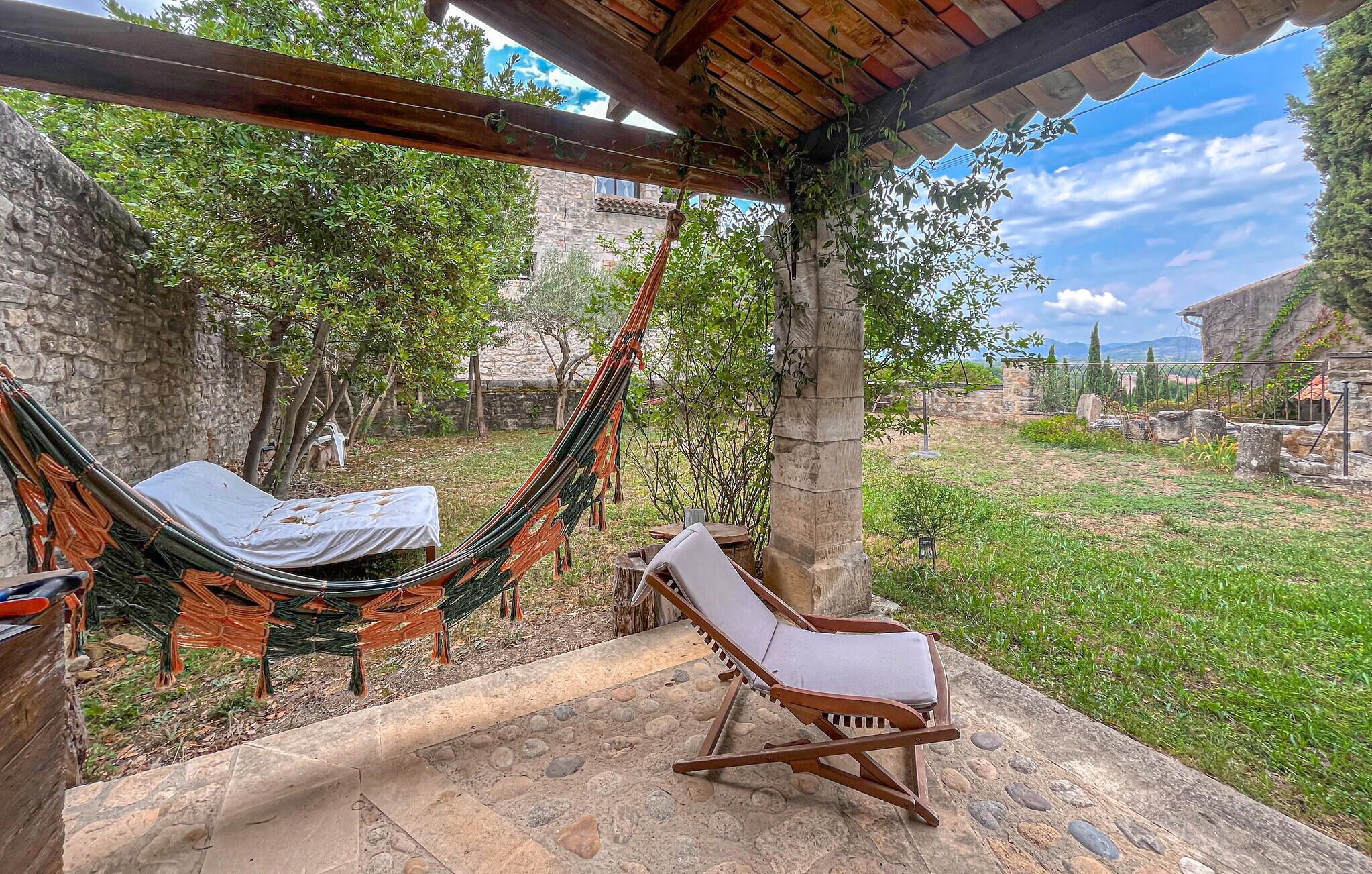 Un hamac et un fauteuil de jardin sous une pergola en bois, dans un jardin aux murs de pierre et à la végétation luxuriante.