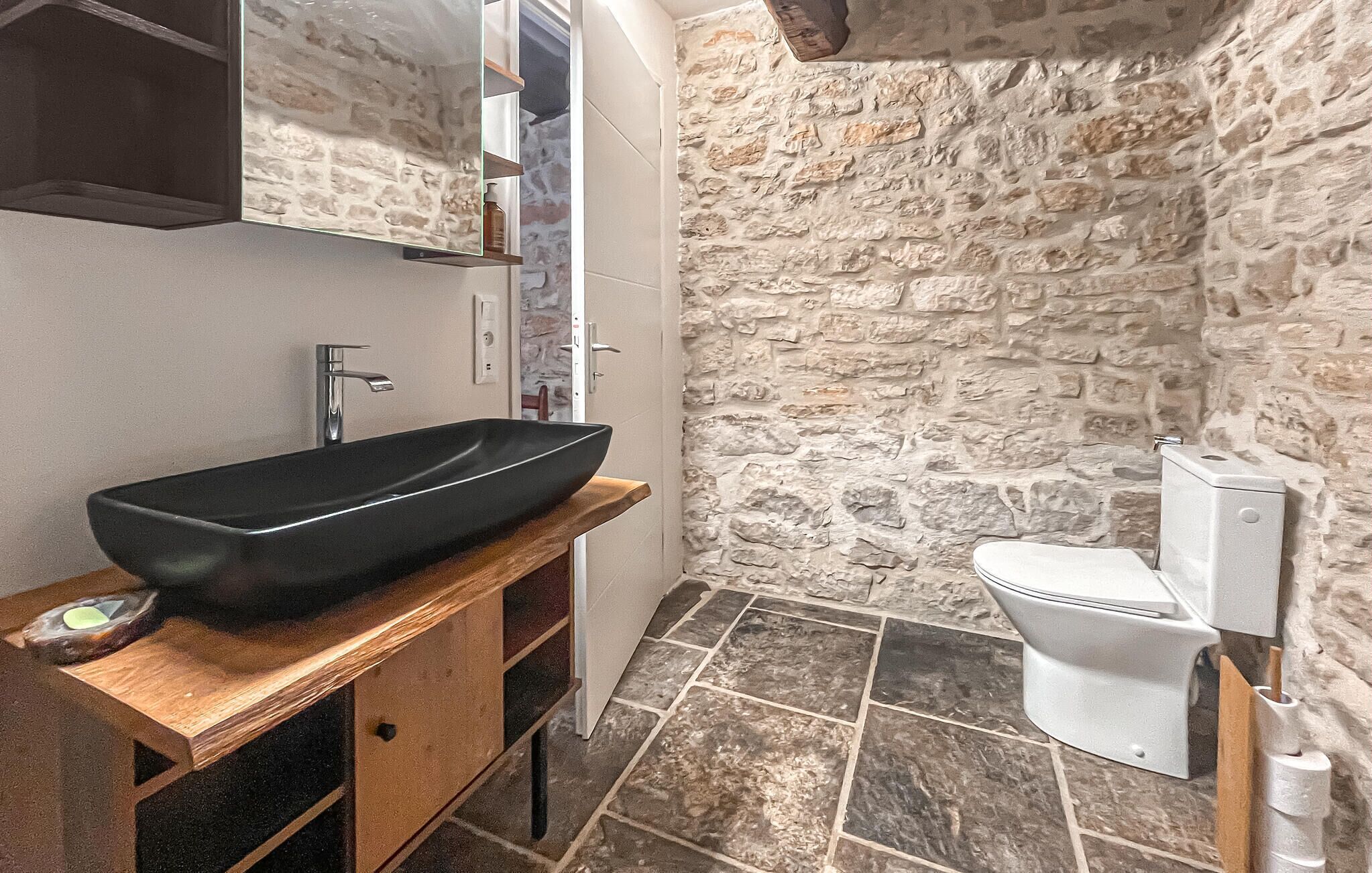 A bathroom with a stone wall, a black vessel sink, a white toilet, and a wooden vanity.