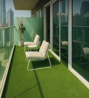 A balcony with artificial grass, two white cushioned chairs, a potted plant, and a glass railing.