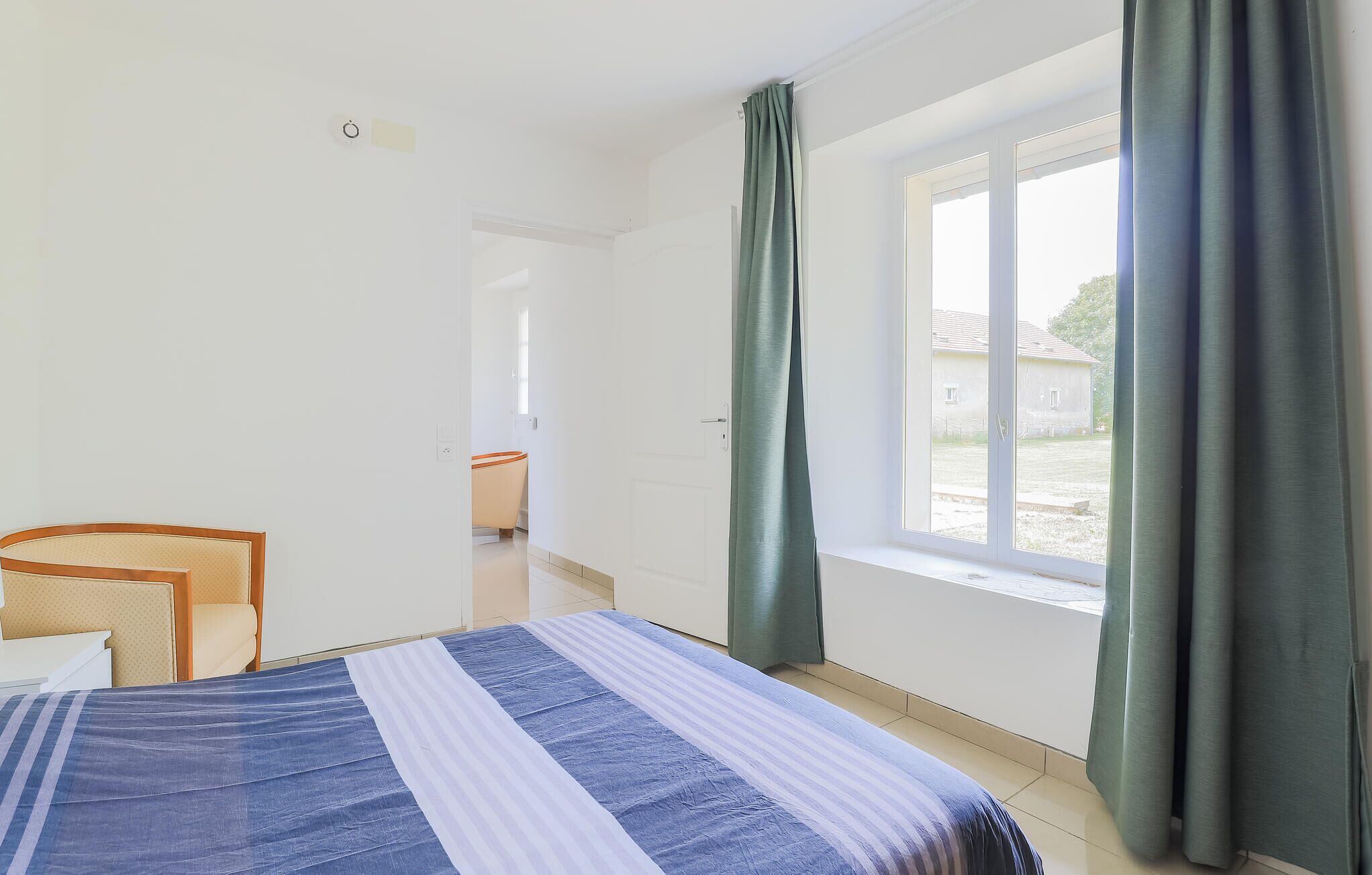 Une chambre à coucher avec un lit, une chaise, une fenêtre avec des rideaux et une vue sur l’extérieur.