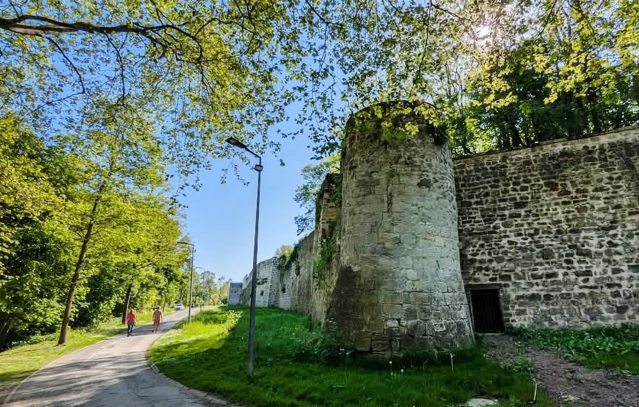 Un mur de pierre surmonté d’une grande tour, un chemin pavé et de la verdure.