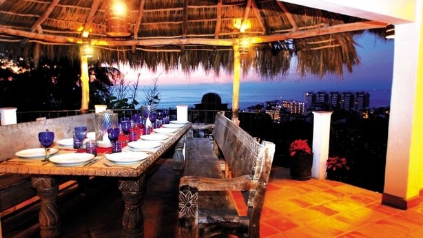 A dining area with a table set for four, a bench, and a view of the ocean at dusk.