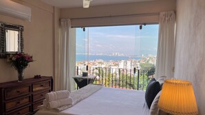 A bedroom with a large bed, a nightstand, a lamp, a dresser, a mirror, and a view of a cityscape and sea.