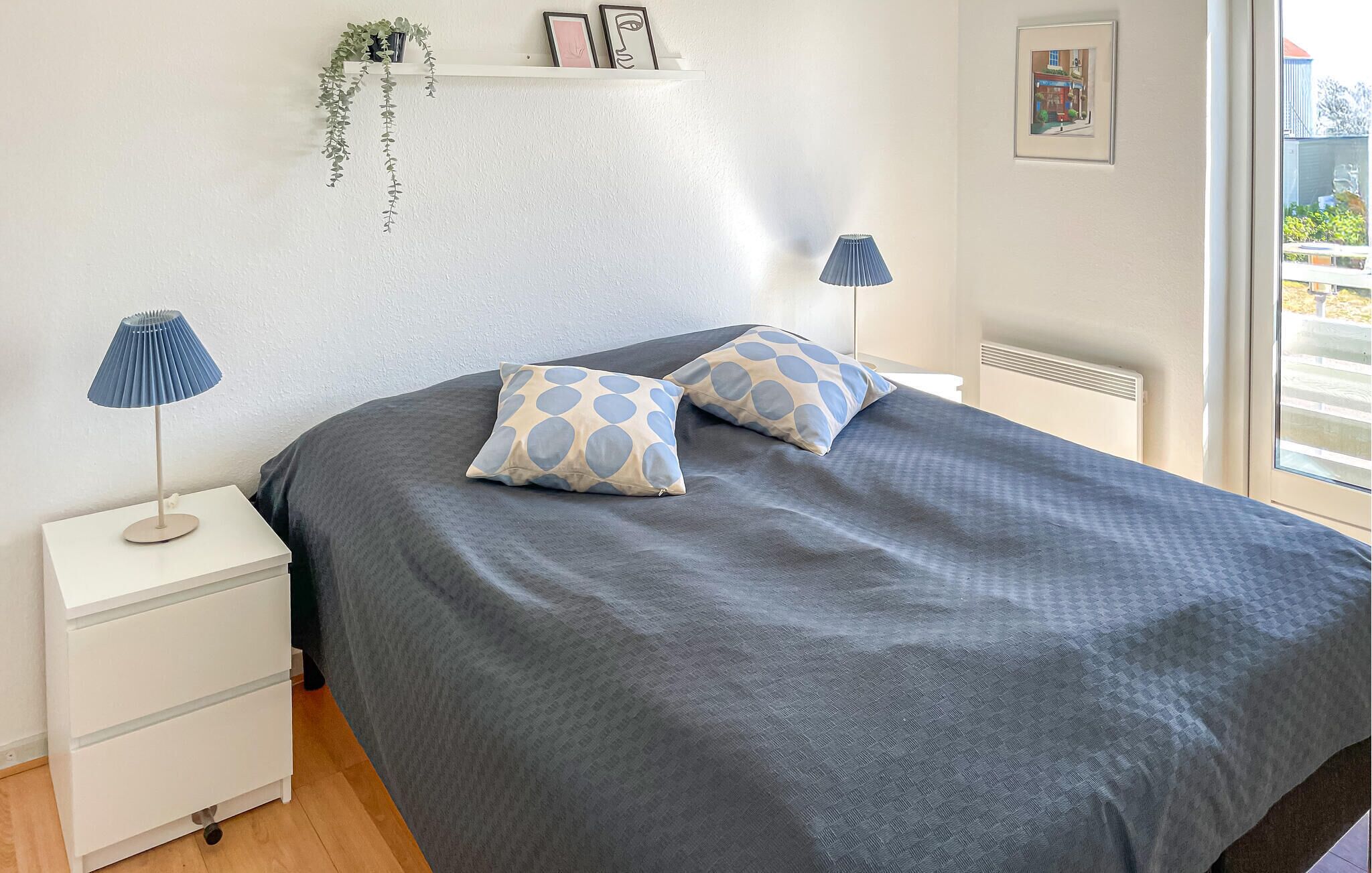 Een slaapkamer met een bed, twee bedlampjes, een nachtkastje, een dressoir, een raam en een wandplank met decoratieve objecten.