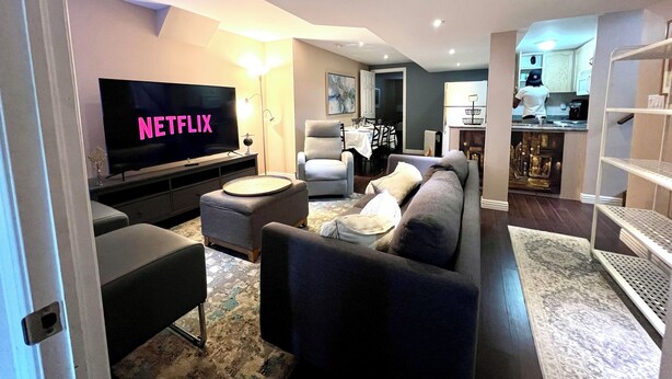 Un salon moderne avec un téléviseur à écran plat affichant le logo « NETFLIX », un canapé gris, une table basse et, au fond, un coin repas.