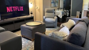 Un salon avec une grande télévision à écran plat affichant le logo « NETFLIX », un fauteuil gris, un canapé gris et, au fond, un coin repas.