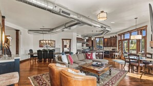 Un salon spacieux avec un grand canapé, des poufs et une table basse. On y trouve également un coin repas avec une table et des chaises. La cuisine est ouverte sur le salon et se distingue par ses meubles en bois et une grande fenêtre.
