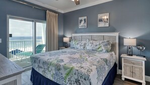 A bedroom with a large bed, two wall-mounted lamps, a ceiling fan, and a view of the sea through a sliding glass door.