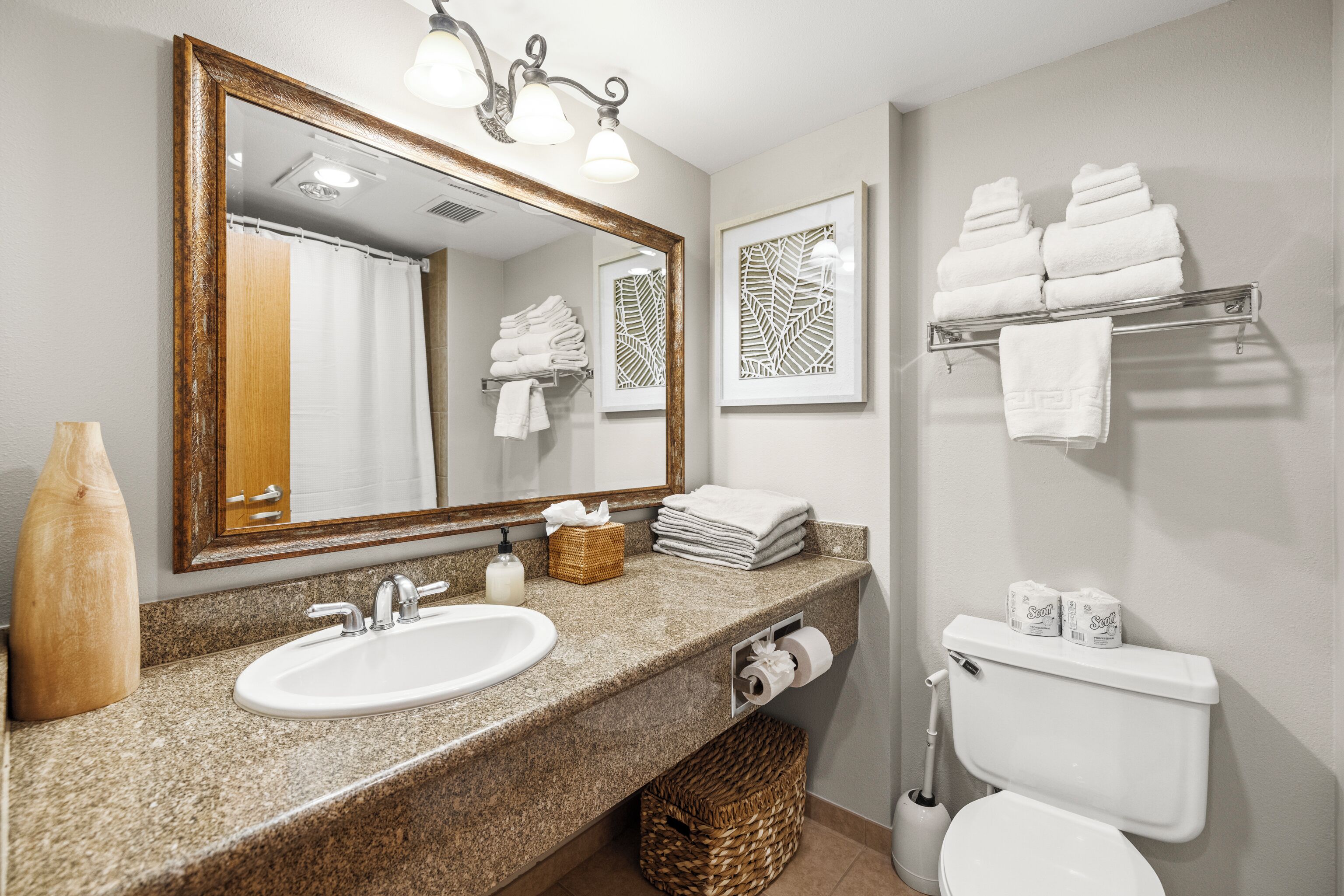 Une salle de bain avec un comptoir en granit, un lavabo blanc, un grand miroir, des toilettes et un porte-serviettes.