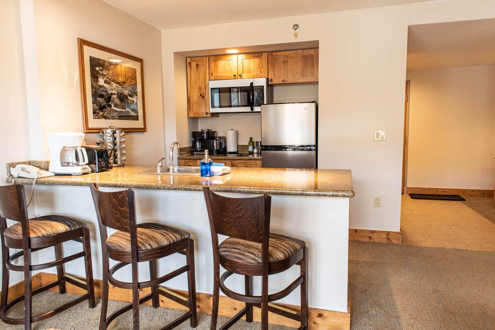 A kitchen with wooden cabinets, a stainless steel refrigerator, a microwave, and a coffee maker. There are three wooden bar stools with cushioned seats.