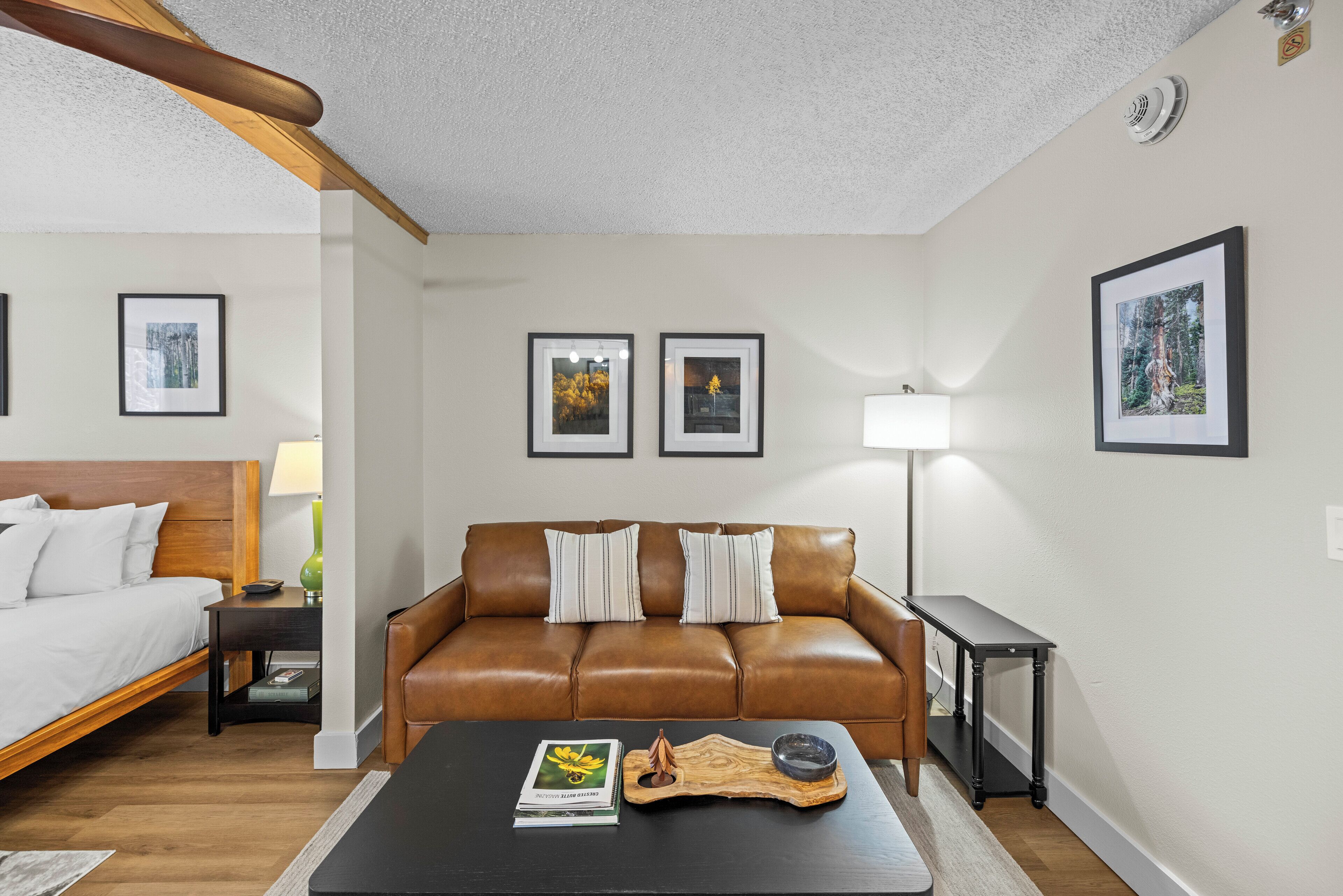 Una habitación de hotel moderna con un sofá de cuero marrón, una mesa de centro de madera, una mesita de noche con lámpara y cuadros enmarcados en las paredes.