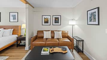 A modern hotel room with a brown leather sofa, a wooden coffee table, a bedside table with a lamp, and framed pictures on the walls.