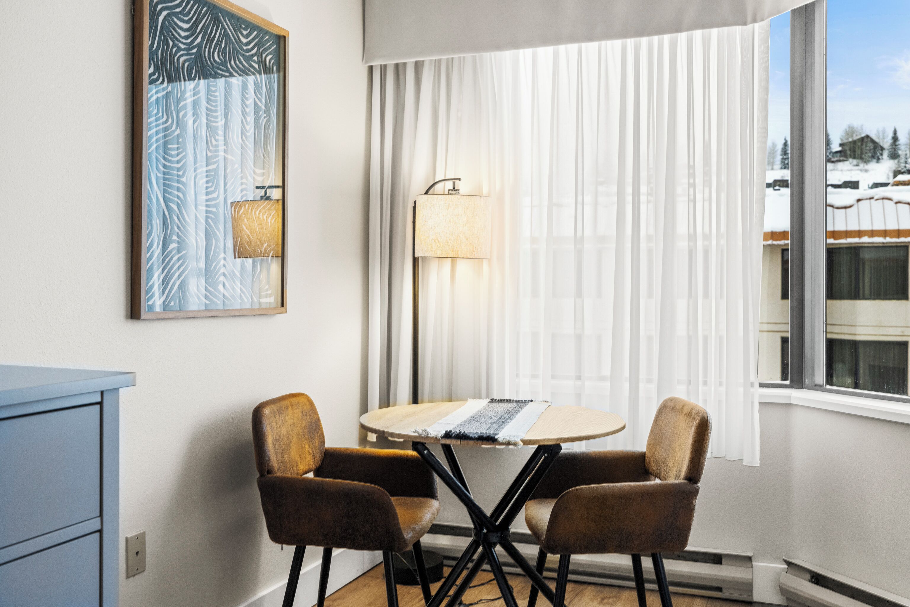 A room with a round table, two chairs, a lamp, a framed artwork, and a window with a view of a snowy landscape.