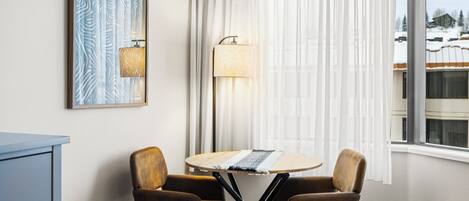 A room with a round table, two chairs, a lamp, a framed artwork, and a window with a view of a snowy landscape.
