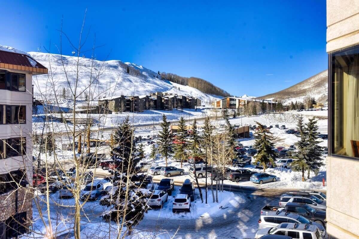 A snow-covered parking lot with cars and trees, buildings with a modern design, and a mountain in the background.