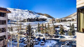 A snow-covered parking lot with cars and trees, buildings with a modern design, and a mountain in the background.