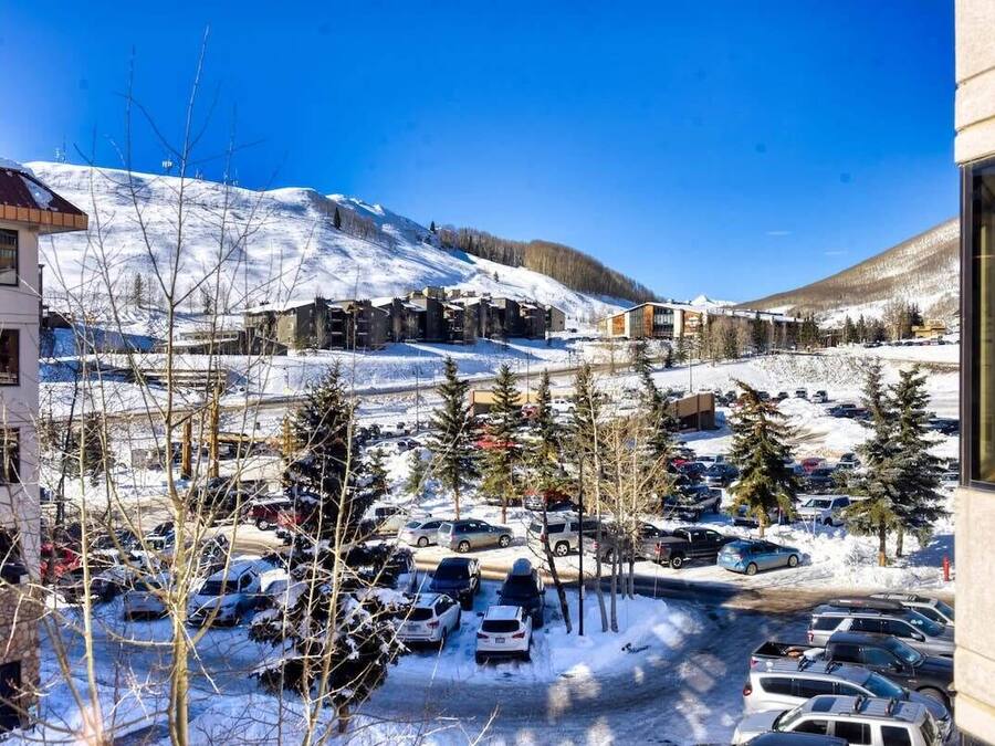 A snow-covered parking lot with cars and trees, buildings with a modern design, and a mountain in the background.
