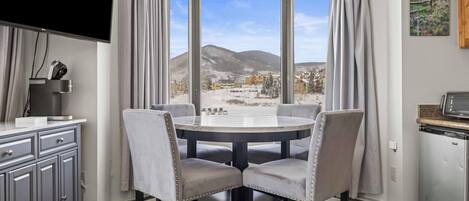 Un comedor con una mesa redonda y cuatro sillas, un televisor montado en la pared y vistas a un paisaje nevado a través de amplios ventanales.