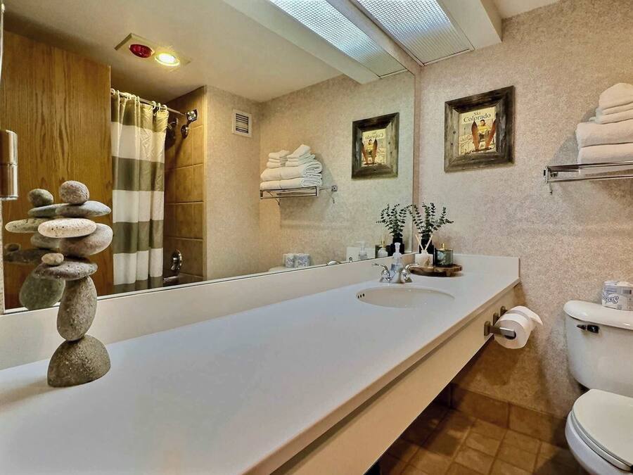 Un baño moderno con un lavamanos alargado, un espejo grande y un inodoro. Hay toallas apiladas en un estante, una planta pequeña en el lavamanos y un cuadro en la pared.