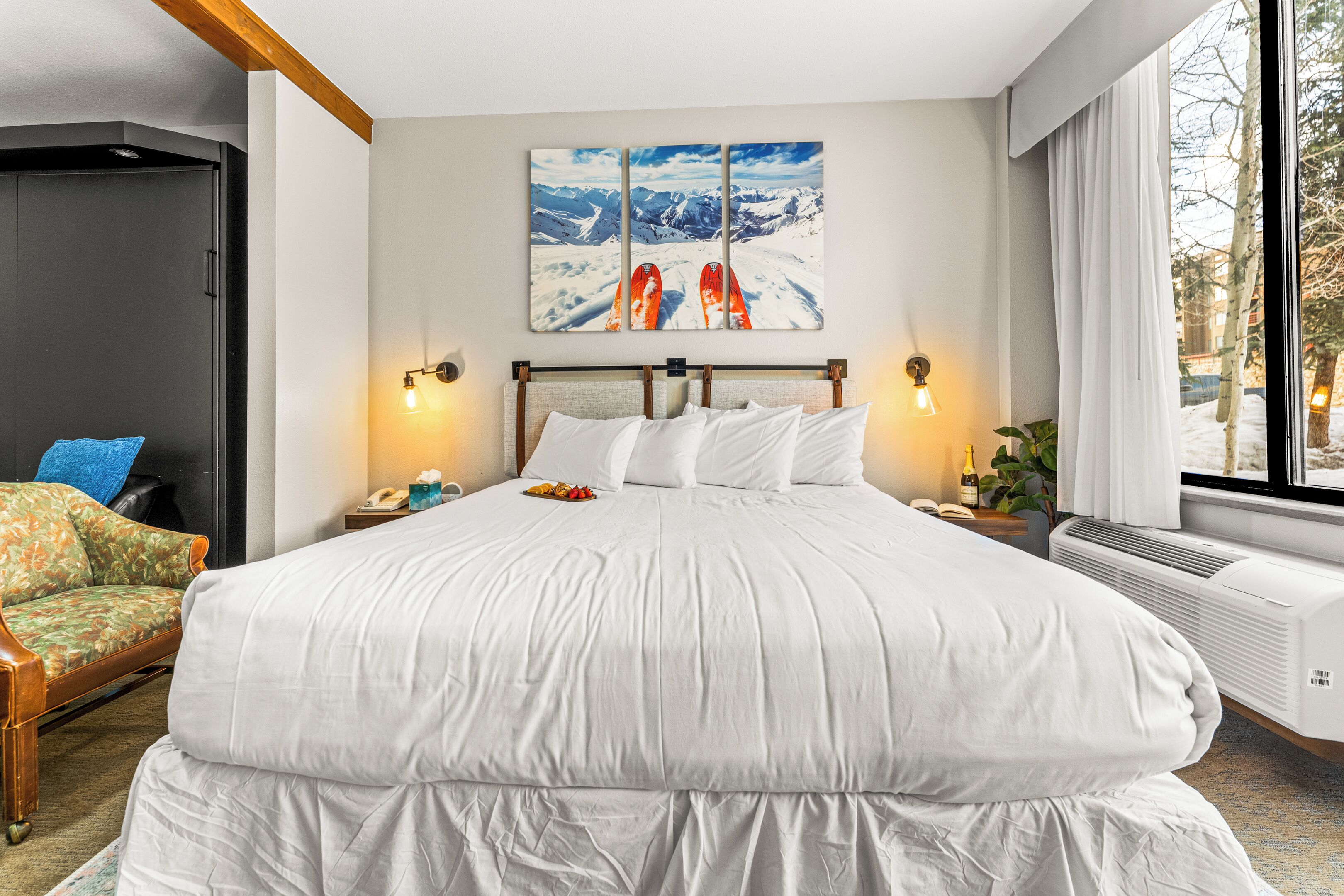 A hotel room with a large bed, a wooden headboard, a nightstand with a lamp, a chair, and a window with a view of mountains.