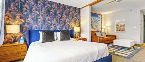 A bedroom with a large bed, a ceiling fan, a nightstand with a lamp, a wooden dresser, and a painting on the wall.