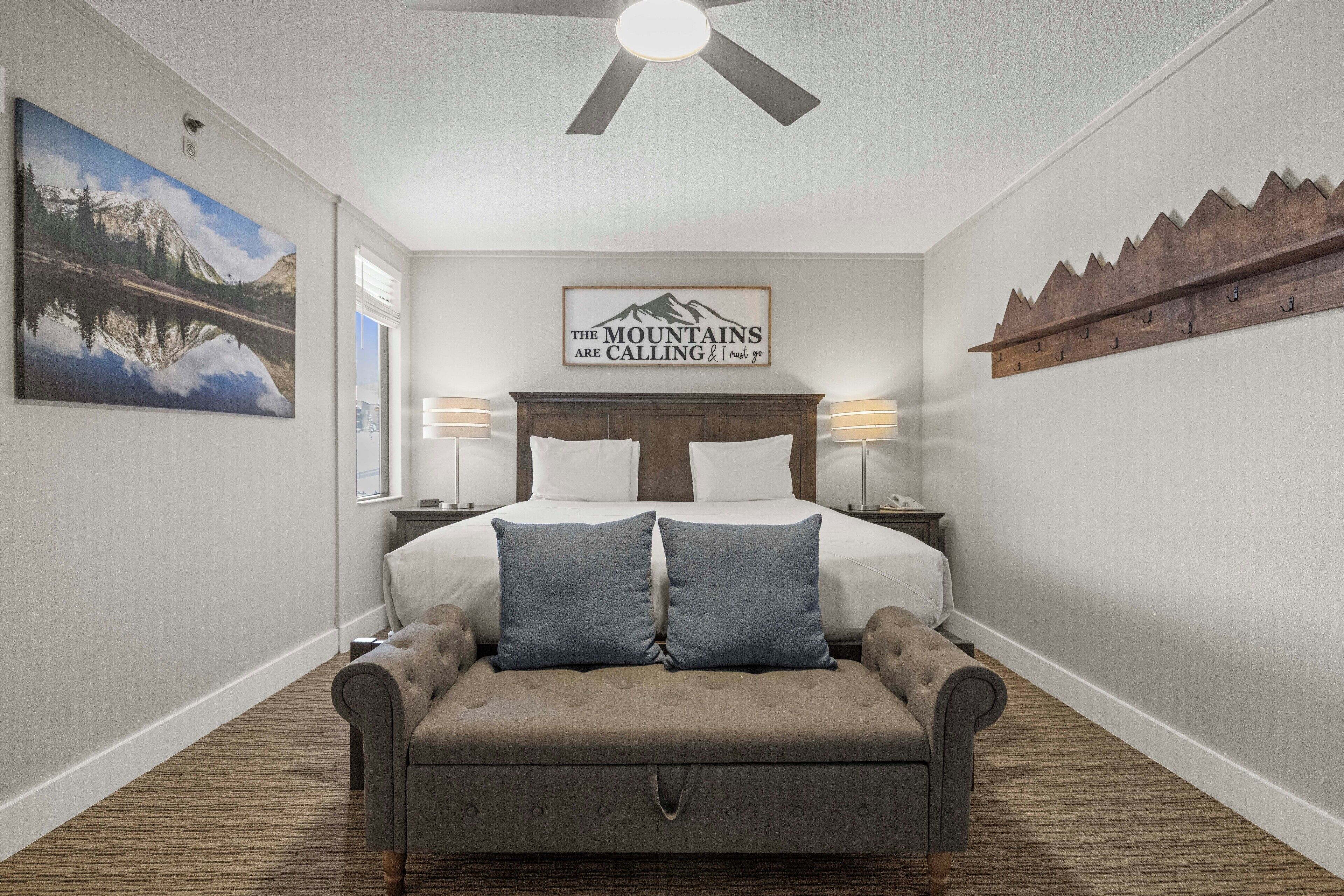 A bedroom with a bed, two bedside lamps, a ceiling fan, a mountain painting, and a wooden shelf with hooks.