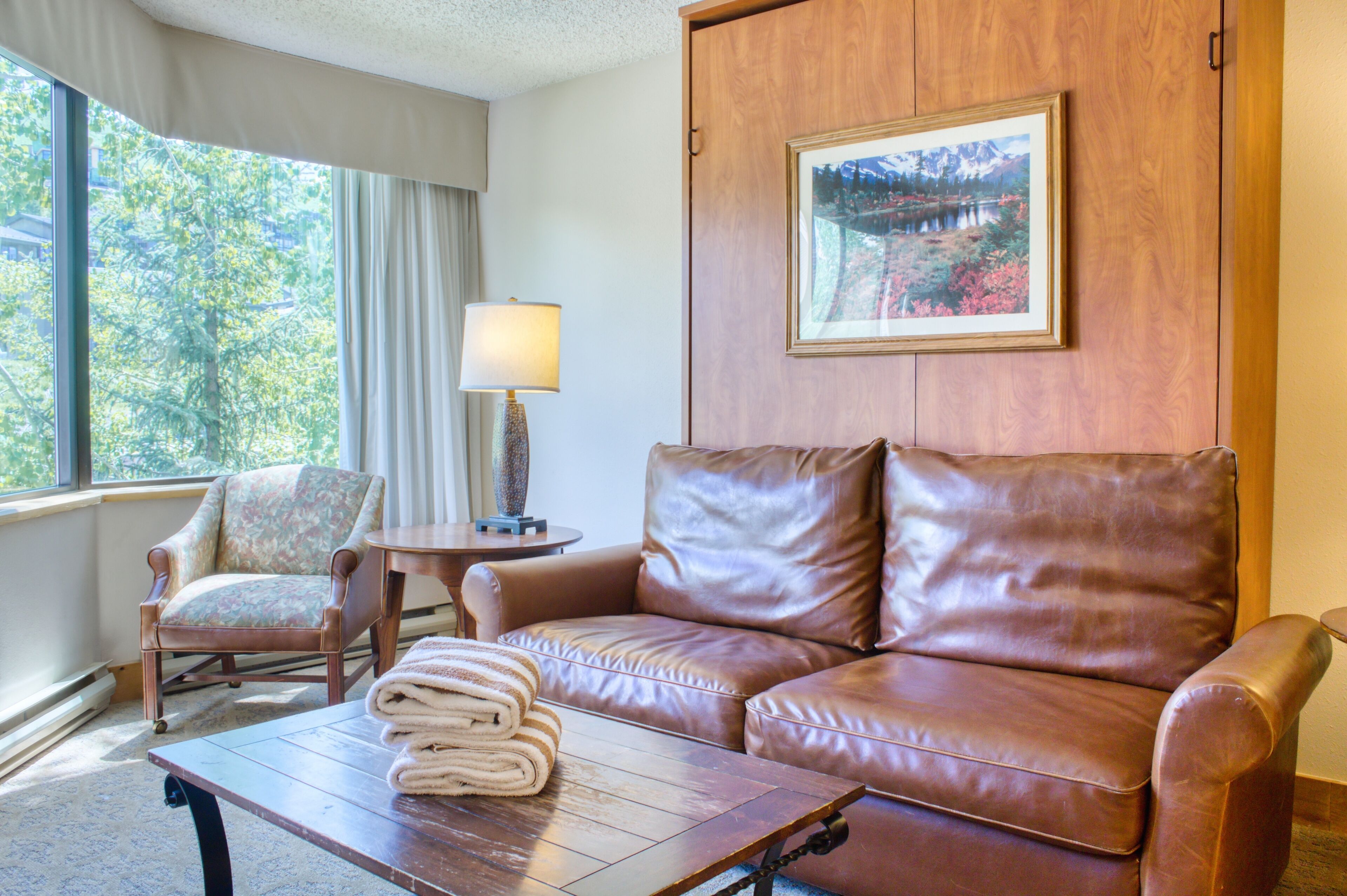 A room with a leather sofa, a wooden armchair, a small table with a lamp, and a framed picture on the wall.