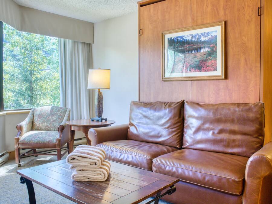 A room with a leather sofa, a wooden armchair, a small table with a lamp, and a framed picture on the wall.