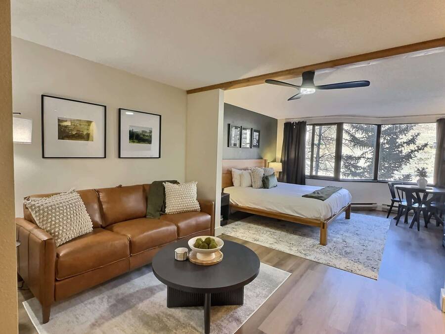 A modern hotel room with a brown leather sofa, a round coffee table, a bed with a wooden frame, and a dining area with a table and chairs.