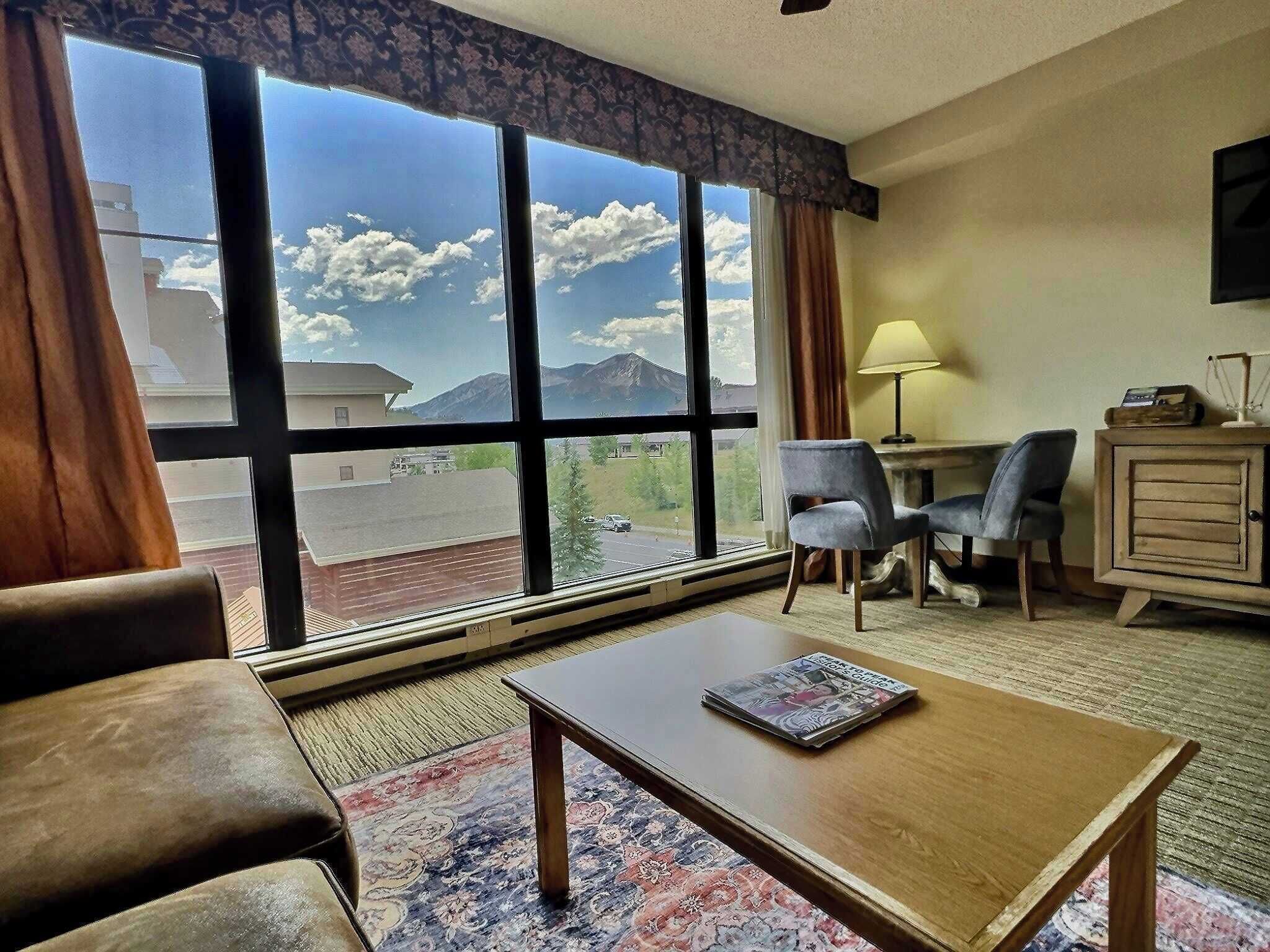 A hotel room with a large window offering a mountain view, a wooden coffee table, a sofa, a desk with a lamp, and a small cabinet.